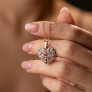 Peut inclure: Collier en or rose avec un pendentif en forme de c&oelig;ur. Le pendentif est incrust&eacute; de petits cristaux transparents et comporte un c&oelig;ur rose plus petit au centre. Le collier est tenu par une personne.