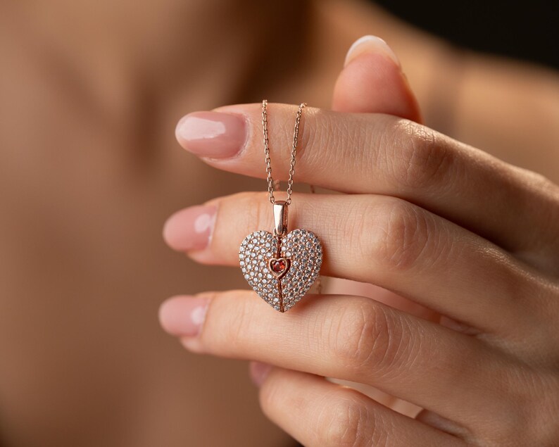 Peut inclure: Collier en or rose avec un pendentif en forme de c&oelig;ur. Le pendentif est incrust&eacute; de petits cristaux transparents et comporte un c&oelig;ur rose plus petit au centre. Le collier est tenu par une personne.