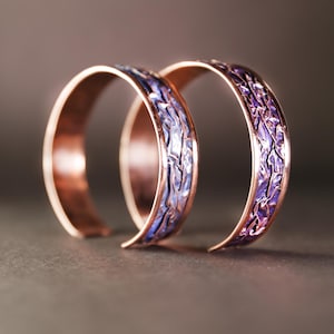Peut inclure: Deux bagues en métal couleur cuivre avec une incrustation texturée violette et bleue. Les bagues sont légèrement incurvées et semblent faites à la main. Le fond est de couleur sombre et neutre, mettant en valeur les bijoux.