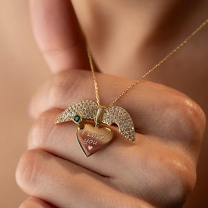 Peut inclure: Un collier dor&eacute; avec un pendentif en forme de c&oelig;ur portant l'inscription "I Love You". Le pendentif est orn&eacute; d'une petite pierre verte et de d&eacute;tails en forme d'ailes sertis de petites pierres claires. Le collier est tenu dans une main.
