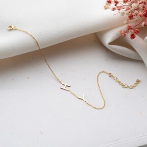 Puede incluir: Un brazalete de cadena dorada con las letras "I" e "Y" colgando de &eacute;l. El brazalete est&aacute; sobre una superficie blanca.