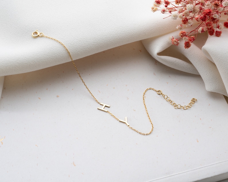 Puede incluir: Un brazalete de cadena dorada con las letras "I" e "Y" colgando de &eacute;l. El brazalete est&aacute; sobre una superficie blanca.