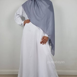 May include: A white abaya dress with long sleeves and a flowing skirt. A gray hijab is draped over the shoulders. The text "pureabayas" is visible on the lower right side of the image.