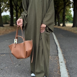 Peut inclure: Une abaya vert olive et un hijab assorti. Un sac fourre-tout en cuir marron clair avec des détails blancs est tenu à la main. La personne porte des baskets blanches. L'arrière-plan est une route bordée d'arbres.