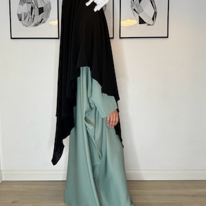Könnte beinhalten: Eine lange, fließende, salbeigrüne Abaya mit einem schwarzen Hijab, der über den Kopf gelegt wird. Die Abaya hat eine lockere, fließende Passform und ist aus einem weichen, seidigen Stoff gefertigt. Die Person trägt weiße Turnschuhe.