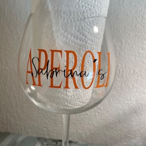 May include: A clear glass wine glass with an orange vinyl sticker that says "Aperol Sabrina's".