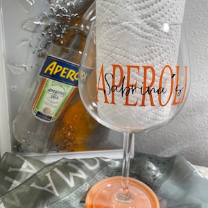 May include: A clear glass with an orange rim and base, decorated with the text "Aperol Sabrina's" in orange and black lettering. The glass is sitting on a light orange coaster.