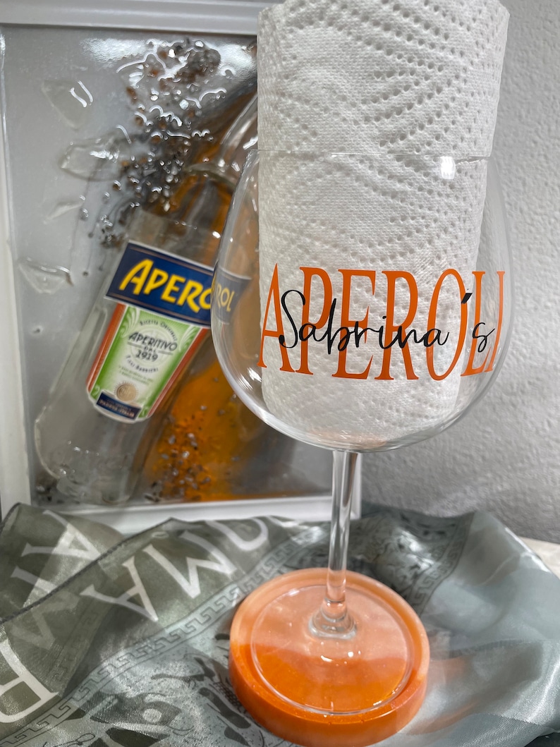 May include: A clear glass with an orange rim and base, decorated with the text "Aperol Sabrina's" in orange and black lettering. The glass is sitting on a light orange coaster.