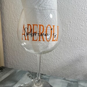 May include: A clear glass wine glass with a stem and a wide bowl. The glass has the word "APEROL" written in orange letters and the words "Sabrina's" written in black cursive letters.