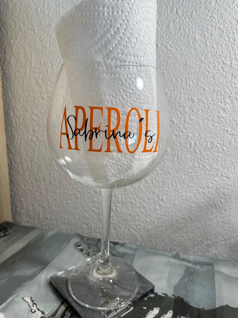 May include: A clear glass wine glass with a stem and a wide bowl. The glass has the word "APEROL" written in orange letters and the words "Sabrina's" written in black cursive letters.