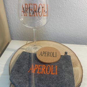 May include: A gray felt pouch with the word "APEROL" in orange lettering, a cork coaster with the word "APEROL" in orange lettering, a clear glass with the word "APEROL" in orange lettering, and a clear glass straw.