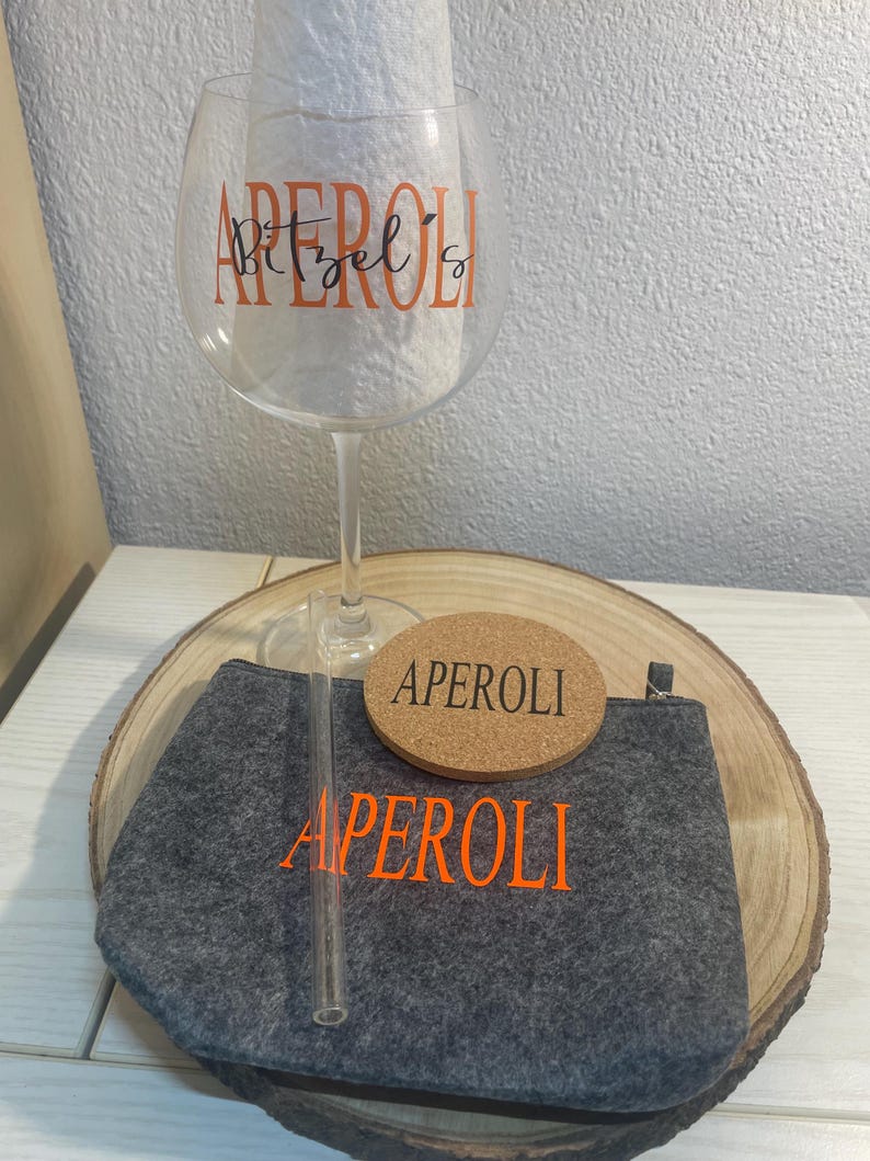 May include: A gray felt pouch with the word "APEROL" in orange lettering, a cork coaster with the word "APEROL" in orange lettering, a clear glass with the word "APEROL" in orange lettering, and a clear glass straw.