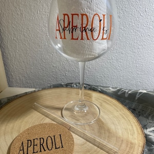 May include: A clear glass with a stem and a wide bowl. The glass has the word "APEROL" written on it in orange letters with the name "Sabrina" written underneath in black letters. A clear glass straw and a cork coaster with the word "APEROL" written on it are also in the image.