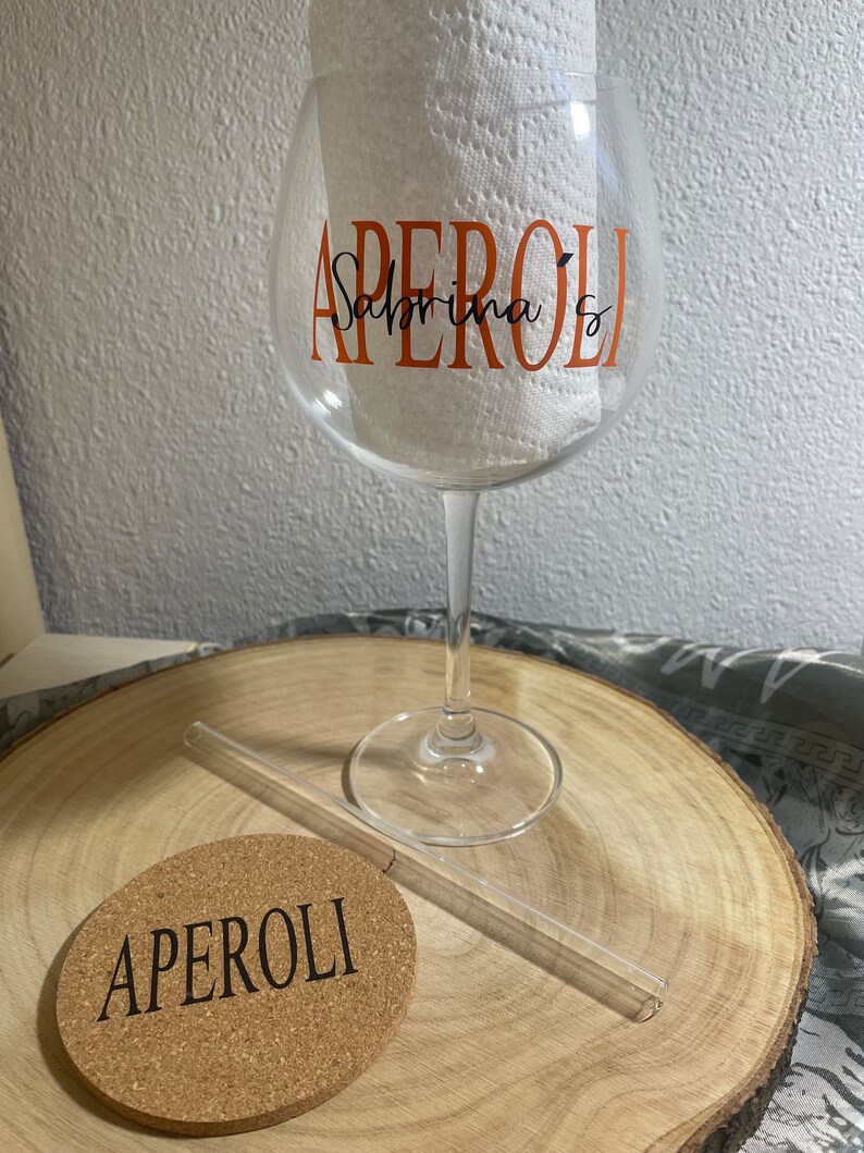 May include: A clear glass with a stem and a wide bowl. The glass has the word "APEROL" written on it in orange letters with the name "Sabrina" written underneath in black letters. A clear glass straw and a cork coaster with the word "APEROL" written on it are also in the image.