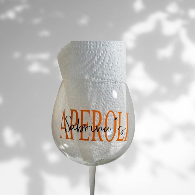 May include: A clear glass with an orange "Aperol Sabrina's" design, partially covered by a white paper towel.