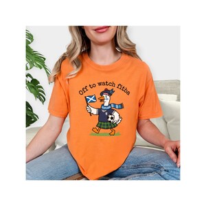 Orange t-shirt lifestyle mockup worn by a woman, featuring a Scottish goose soccer supporter graphic with “Off to watch fitba” text and Scotland details.