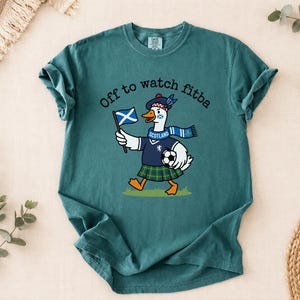 Blue Spruce Comfort Colors style t-shirt flat lay featuring a funny Scotland goose football fan design with “Off to watch fitba” text and soccer ball.