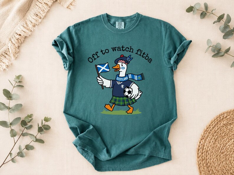 Blue Spruce Comfort Colors style t-shirt flat lay featuring a funny Scotland goose football fan design with “Off to watch fitba” text and soccer ball.