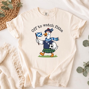 Close-up Ivory t-shirt mockup showing the Scotland goose football fan design, “Off to watch fitba” text, Scottish flag, kilt, scarf, and soccer ball.