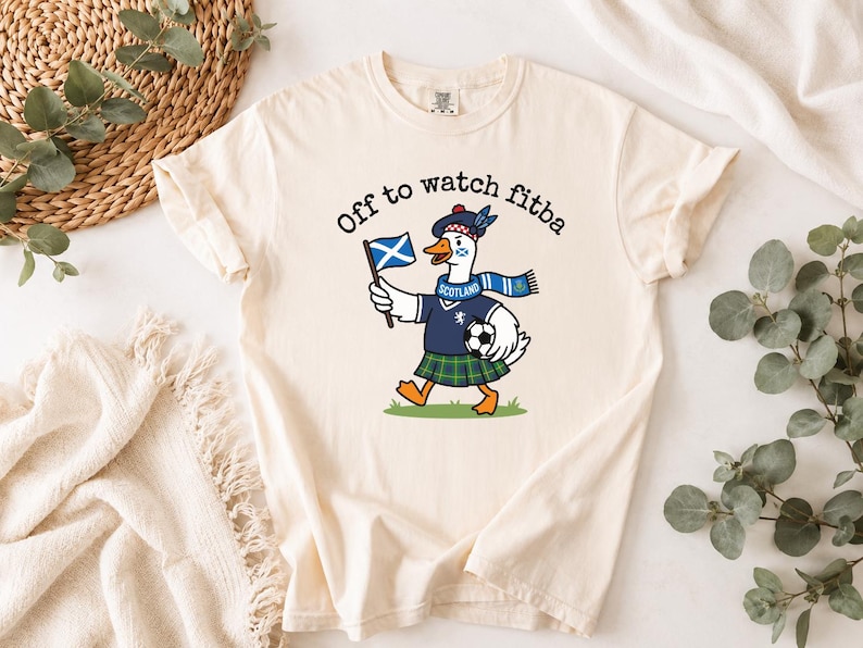 Close-up Ivory t-shirt mockup showing the Scotland goose football fan design, “Off to watch fitba” text, Scottish flag, kilt, scarf, and soccer ball.