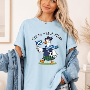 Chambray blue t-shirt mockup worn by a woman, featuring a Scottish goose football fan design with “Off to watch fitba” text and Scotland scarf graphic.