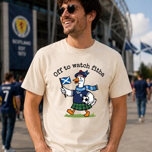 Ivory Comfort Colors style t-shirt flat lay featuring a Scotland goose soccer fan graphic with “Off to watch fitba” text, styled with neutral boho decor.