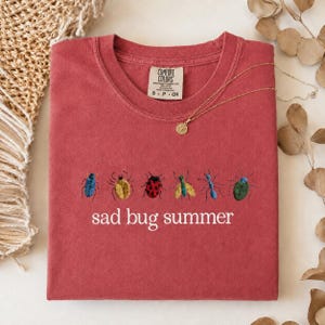 May include: A brick-red t-shirt featuring embroidered insects in various colours and the words "sad bug summer." The top has a round neck and short sleeves. A gold necklace with a pendant is draped across the shirt.