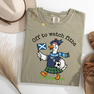 Sandstone green folded t-shirt flat lay with Scotland goose soccer fan graphic, “Off to watch fitba” text, Scottish flag, kilt, and football design.