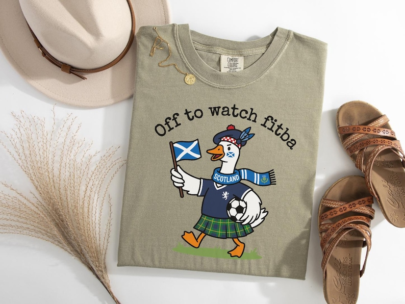 Sandstone green folded t-shirt flat lay with Scotland goose soccer fan graphic, “Off to watch fitba” text, Scottish flag, kilt, and football design.