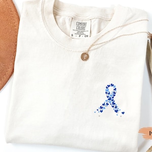 May include: Ivory-colored t-shirt with a blue heart-shaped ribbon design. The ribbon is made of various shades of blue hearts. A gold necklace with a pendant is draped on the shirt. The shirt is labeled "Comfort Colors".