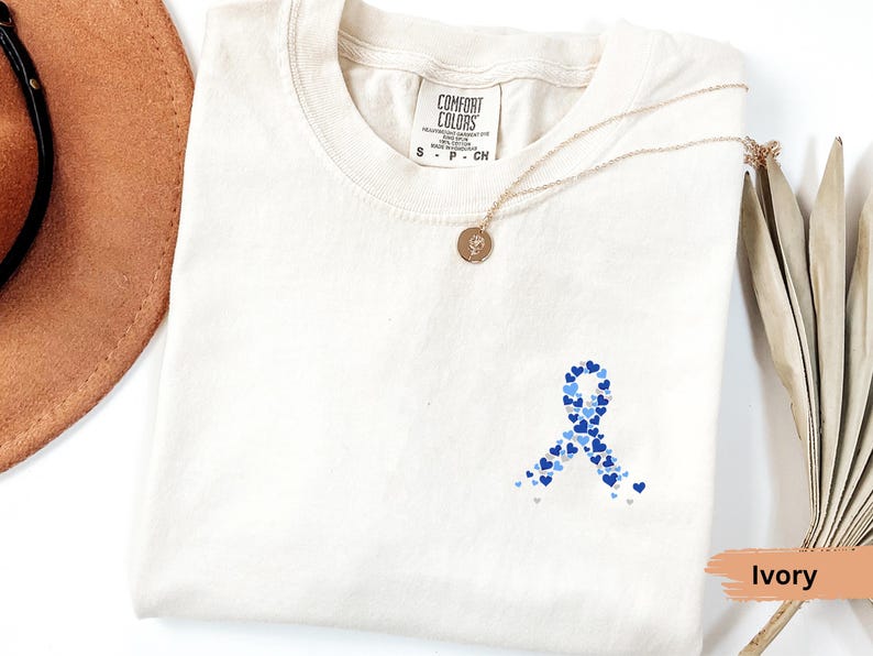 May include: Ivory-colored t-shirt with a blue heart-shaped ribbon design. The ribbon is made of various shades of blue hearts. A gold necklace with a pendant is draped on the shirt. The shirt is labeled "Comfort Colors".