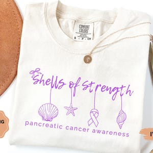May include: Ivory-colored t-shirt with the text "Shells of Strength" in purple, along with seashell, starfish, ribbon, and conch graphics. The phrase "pancreatic cancer awareness" is printed below the graphics. A gold necklace is draped on the shirt.