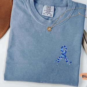 May include: A blue jean colored t-shirt with a small embroidered ribbon made of blue hearts. A gold necklace with a pendant rests on the shirt. The shirt has a Comfort Colors tag. A brown hat and dried plant are in the background.