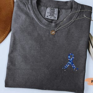 May include: A dark gray t-shirt with a blue ribbon design made of hearts. The shirt has a crew neck and short sleeves. A gold necklace with a pendant is on the shirt. The shirt is labeled "Comfort Colors".