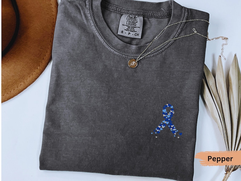 May include: A dark gray t-shirt with a blue ribbon design made of hearts. The shirt has a crew neck and short sleeves. A gold necklace with a pendant is on the shirt. The shirt is labeled "Comfort Colors".