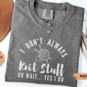 May include: A gray Comfort Colors t-shirt with the text "I DON'T ALWAYS Knit Stuff OH WAIT... YES I DO" in white. The shirt features a graphic of yarn and knitting needles. A gold necklace rests on the shirt.