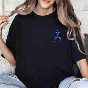 May include: Black t-shirt featuring a blue ribbon design on the chest. The ribbon is made of small blue and white elements. The shirt is a solid black color and has short sleeves. The shirt is made of a soft material.