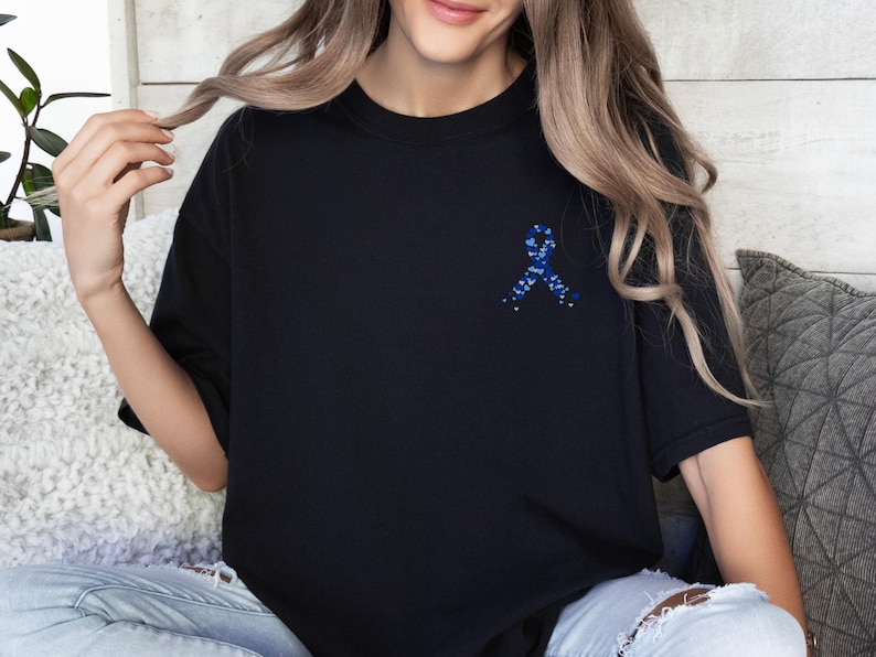 May include: Black t-shirt featuring a blue ribbon design on the chest. The ribbon is made of small blue and white elements. The shirt is a solid black color and has short sleeves. The shirt is made of a soft material.