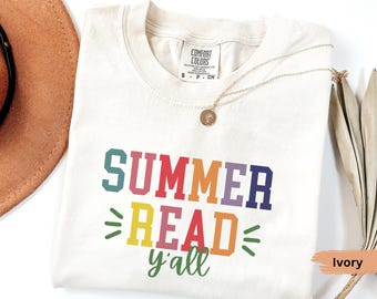 May include: Ivory-colored t-shirt with the text "SUMMER READ y'all" in colorful block letters. The shirt has a crew neck and short sleeves. A gold necklace with a pendant is draped on the shirt.