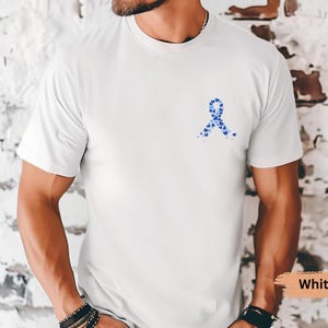 May include: White t-shirt featuring a blue ribbon design made of small hearts. The shirt is a solid white color and has a crew neck. The design is located on the left chest area of the shirt.