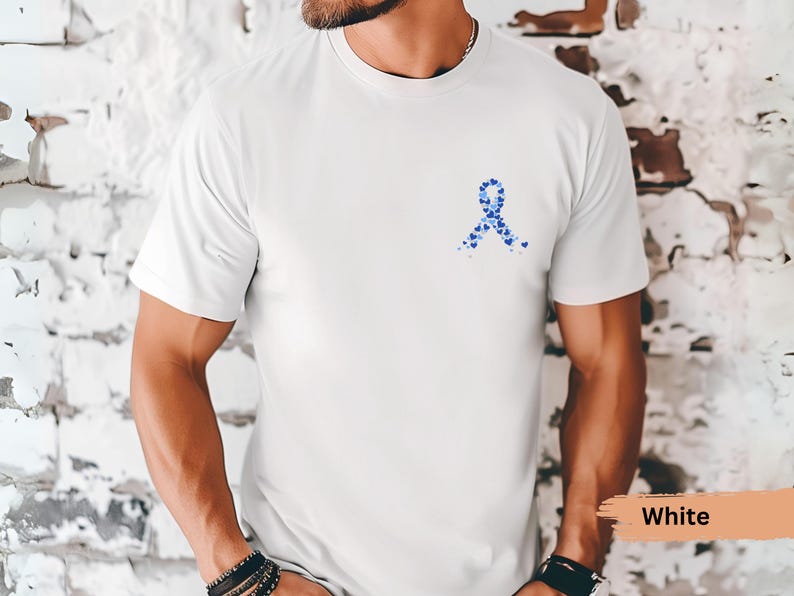 May include: White t-shirt featuring a blue ribbon design made of small hearts. The shirt is a solid white color and has a crew neck. The design is located on the left chest area of the shirt.