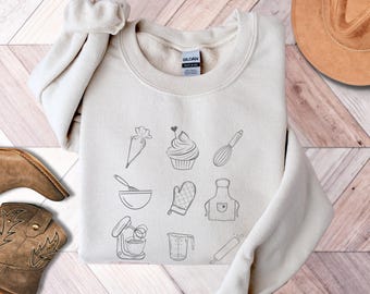 May include: A cream-colored sweatshirt with a graphic design of various baking tools and ingredients, including a whisk, a rolling pin, a cupcake, a measuring cup, a mixing bowl, a kitchen mitt, an apron, and a piping bag.
