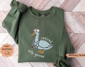 Puede incluir: Sudadera verde militar con un dibujo de un ganso con gorro de enfermera y estetoscopio. El ganso tiene las palabras "retired silly goose" impresas en la parte delantera. La sudadera está doblada sobre una superficie de madera.