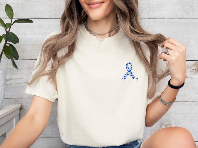 May include: Cream-colored short-sleeved t-shirt featuring a blue ribbon design made of small blue hearts. The shirt is made of a soft material and has a classic crew neck.