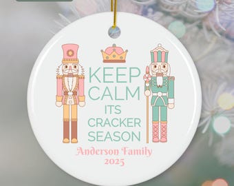 May include: A white ceramic Christmas ornament with a gold string. The ornament features two nutcracker figures and the text "KEEP CALM ITS CRACKER SEASON" with "Anderson Family 2025" below. The ornament is 2.99 inches in diameter.