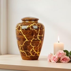 May include: A brown urn with a lid, featuring a gold crackle design. A lit candle and pink roses are next to the urn. The urn is on a wooden surface.
