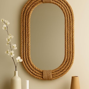 May include: Oval mirror with a natural rope frame. The mirror is surrounded by multiple layers of thick, twisted rope, creating a textured border. The frame is secured with wrapped rope at the top and bottom. A white vase with flowers and a wooden vase are on a wooden surface.