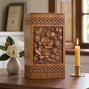 Op de afbeelding: Houten urn met gesneden bloemen- en vlinderdetails. De rechthoekige doos heeft een gesneden bloemenontwerp op het voorpaneel, met Keltische knoopwerkranden. Een brandende kaars in een gouden houder staat in de buurt, met een vaas met witte bloemen.