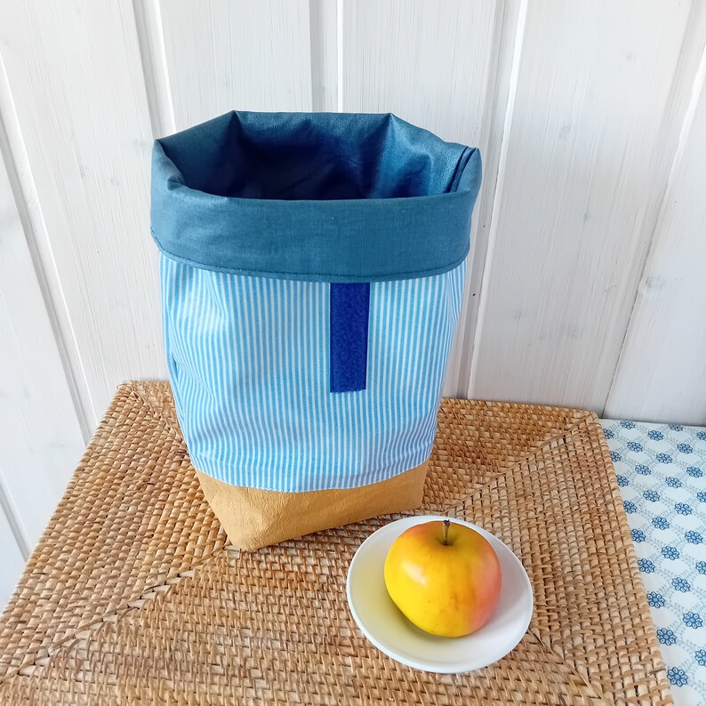 May include: A blue and white striped fabric storage bin with a blue fabric top and a brown bottom. The bin is sitting on a woven wicker mat with a yellow apple on a white plate.