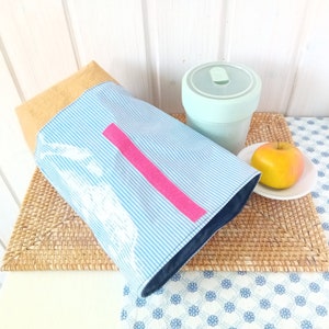 May include: A blue and white striped lunch bag with a pink stripe and a white design. The bag is sitting on a woven placemat next to a light green reusable cup and a yellow apple.